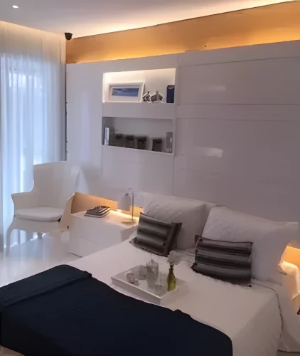 Quarto moderno e minimalista com paredes brancas e iluminação indireta. A cama, de casal, está arrumada com roupas de cama brancas, duas almofadas grandes e duas almofadas menores em tons escuros. Sobre a cama há uma bandeja com garrafa e copos. Ao lado, um criado-mudo branco com luminária de mesa e alguns livros. No fundo, há uma estante embutida com prateleiras iluminadas, exibindo quadros, objetos decorativos e potes. Uma poltrona branca fica próxima a uma cortina translúcida que cobre uma janela grande, permitindo entrada de luz natural.